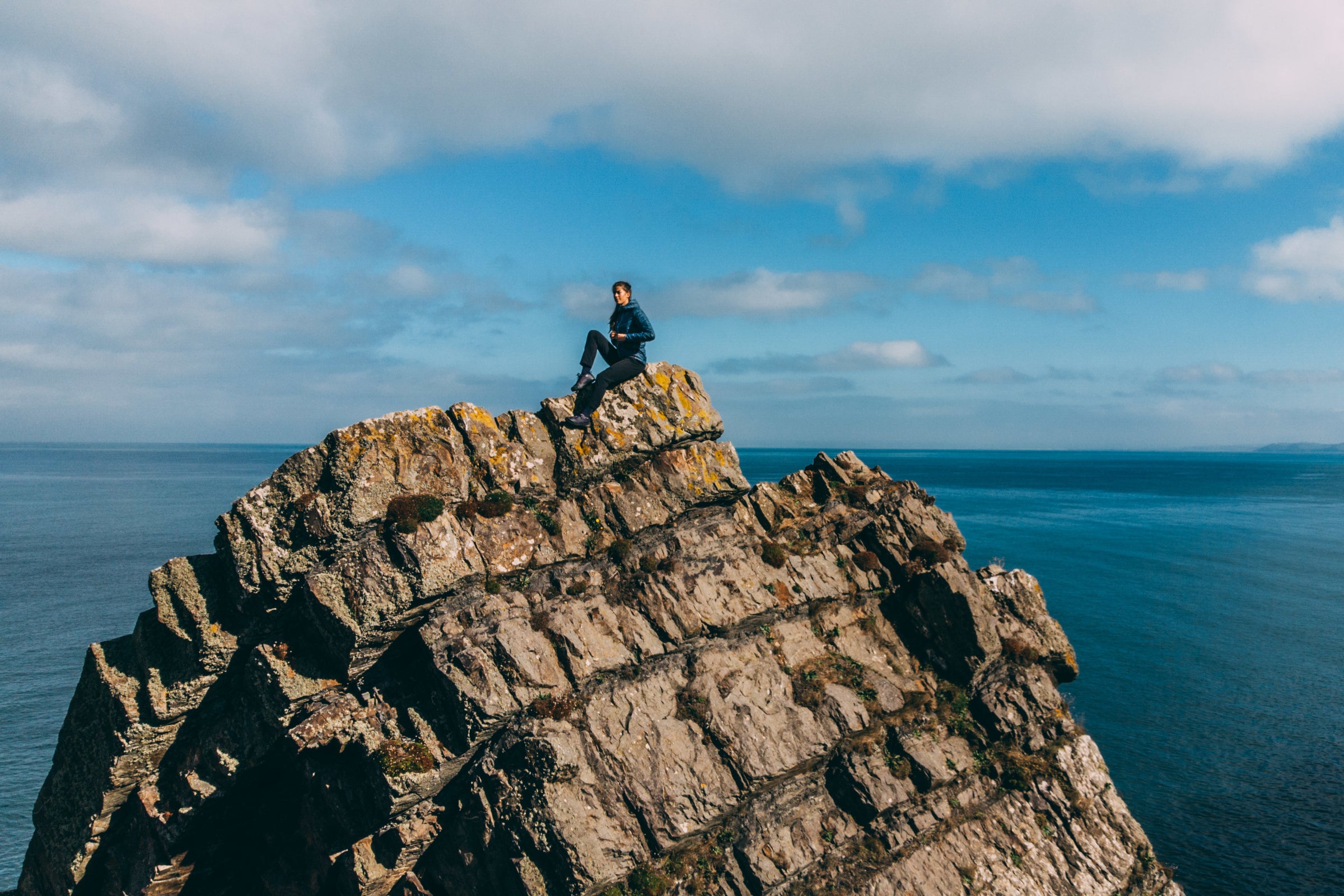 A person sitting on top of a rocky cliff overlooking the ocean under a clear sky